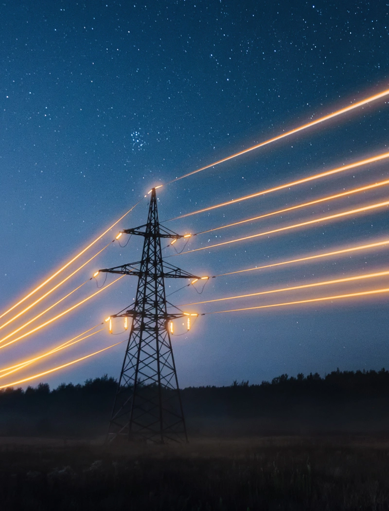 A photograph of a tower carrying high tension power lines, against a starlit night sky. The power lines, and the spots where they make contact with the tower, are illuminated.